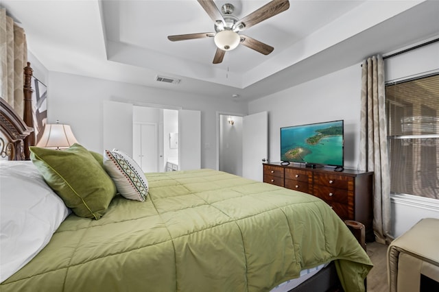 carpeted bedroom featuring visible vents, a raised ceiling, ensuite bathroom, and a ceiling fan