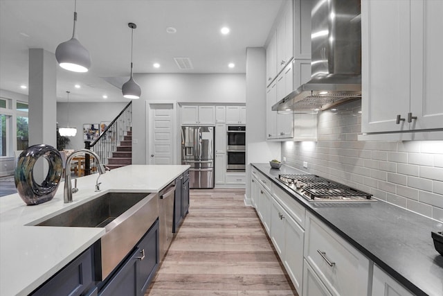 kitchen with light wood-type flooring, wall chimney exhaust hood, backsplash, hanging light fixtures, and appliances with stainless steel finishes