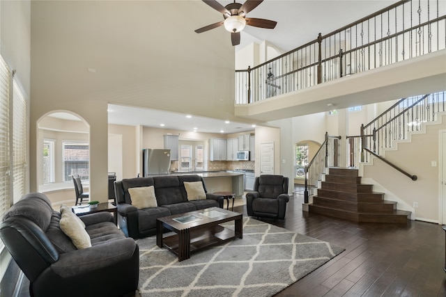 living room with arched walkways, a high ceiling, a ceiling fan, stairs, and dark wood-type flooring