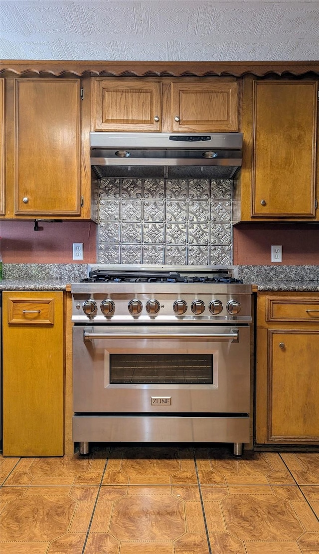 kitchen with high end stainless steel range, dark countertops, decorative backsplash, exhaust hood, and brown cabinetry