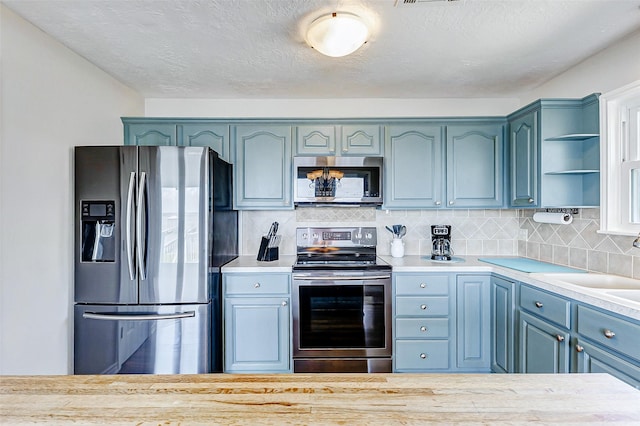 kitchen with open shelves, blue cabinets, light countertops, stainless steel appliances, and backsplash