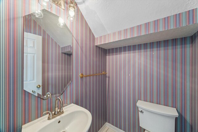half bathroom featuring a sink, toilet, a textured ceiling, and wallpapered walls