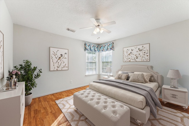 bedroom with baseboards, light wood-type flooring, a textured ceiling, visible vents, and ceiling fan