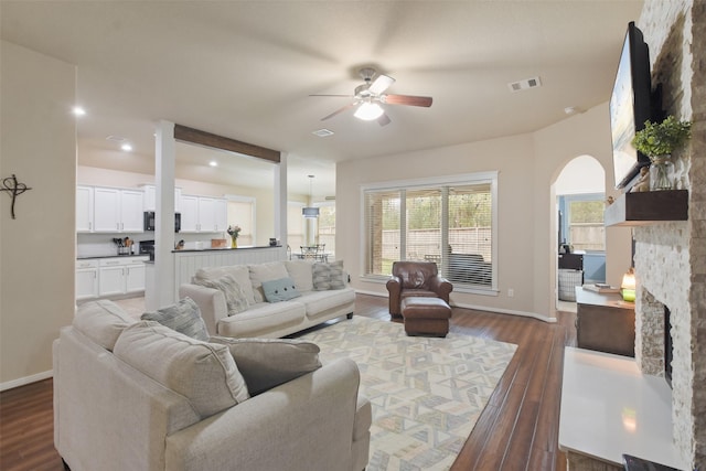 living area featuring a ceiling fan, a stone fireplace, visible vents, wood finished floors, and baseboards
