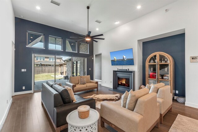 living room with a high ceiling, a warm lit fireplace, wood finished floors, visible vents, and a ceiling fan