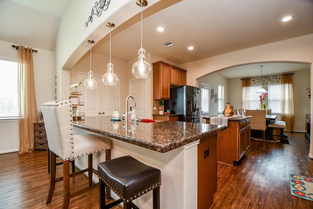 kitchen featuring black refrigerator with ice dispenser, brown cabinets, arched walkways, a center island with sink, and dark wood-style flooring