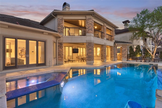 back of house at dusk with a chimney, a balcony, a patio area, stone siding, and a pool with connected hot tub