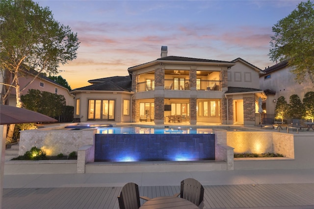 back of property at dusk featuring a patio area, a balcony, an outdoor pool, stone siding, and an outdoor hot tub
