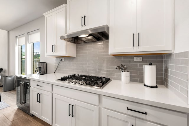 kitchen with under cabinet range hood, wine cooler, stainless steel gas cooktop, tasteful backsplash, and white cabinetry