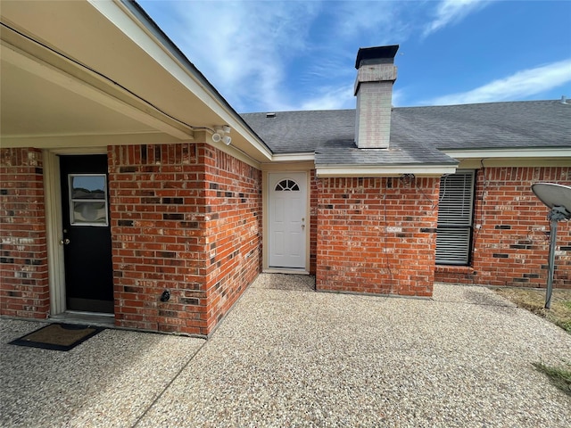 view of exterior entry featuring a chimney, brick siding, a patio, and a shingled roof
