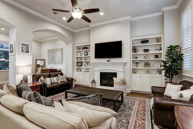 living area with ornamental molding, dark wood finished floors, ceiling fan, arched walkways, and a glass covered fireplace