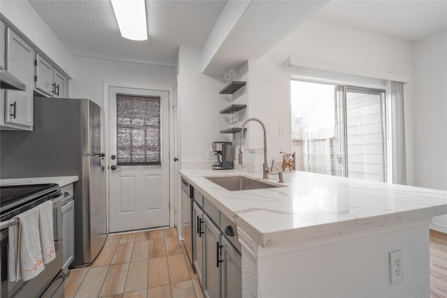 kitchen with a sink, light stone counters, gray cabinets, open shelves, and appliances with stainless steel finishes