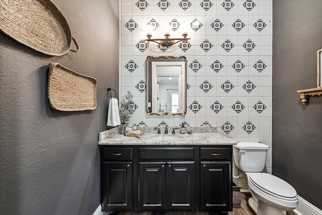 half bath with toilet, a textured wall, baseboards, and vanity