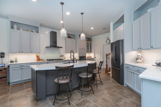 kitchen with glass insert cabinets, an island with sink, wall chimney range hood, a sink, and appliances with stainless steel finishes