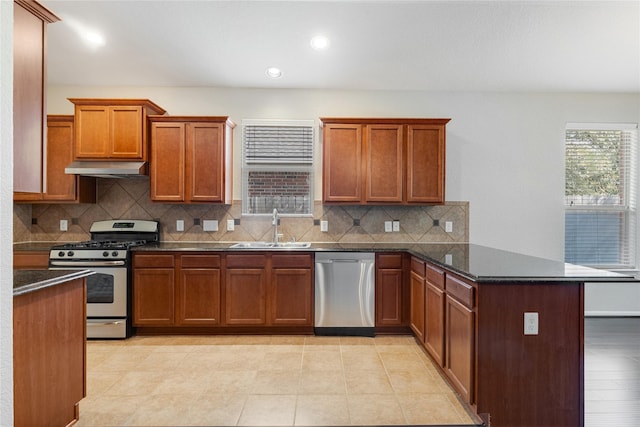 kitchen with a peninsula, a sink, brown cabinetry, under cabinet range hood, and appliances with stainless steel finishes