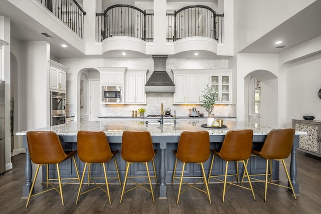 kitchen with a towering ceiling, a large island with sink, premium range hood, stainless steel appliances, and dark wood-type flooring