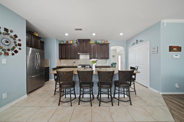 kitchen with arched walkways, dark brown cabinetry, backsplash, stainless steel refrigerator, and visible vents