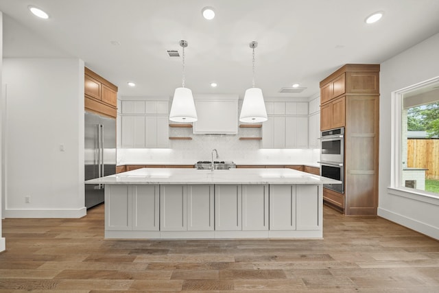 kitchen featuring open shelves, a kitchen island with sink, a sink, backsplash, and stainless steel appliances