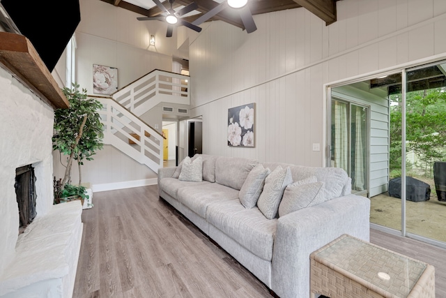 living area with beam ceiling, a fireplace with raised hearth, stairs, a ceiling fan, and wood finished floors