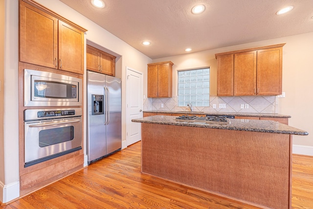 kitchen with light wood finished floors, a center island, dark stone counters, decorative backsplash, and appliances with stainless steel finishes