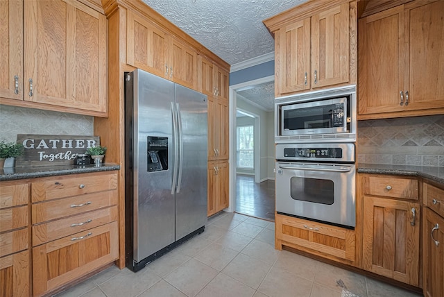 kitchen featuring appliances with stainless steel finishes, ornamental molding, light tile patterned flooring, tasteful backsplash, and dark stone countertops