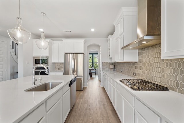 kitchen with arched walkways, a sink, light wood-type flooring, wall chimney range hood, and appliances with stainless steel finishes