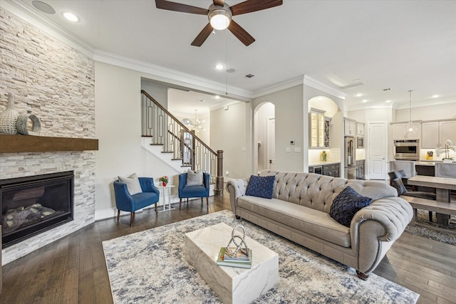 living room featuring dark wood-style flooring, a fireplace, stairway, ceiling fan with notable chandelier, and ornamental molding