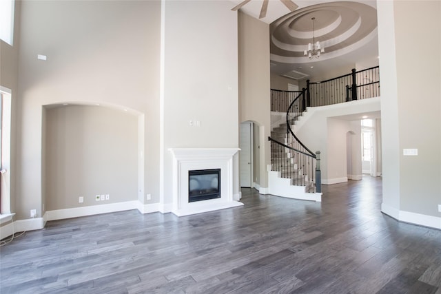 unfurnished living room with stairway, a high ceiling, wood finished floors, a glass covered fireplace, and ceiling fan with notable chandelier