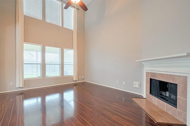 unfurnished living room with dark wood-style floors, a tiled fireplace, a towering ceiling, baseboards, and a ceiling fan