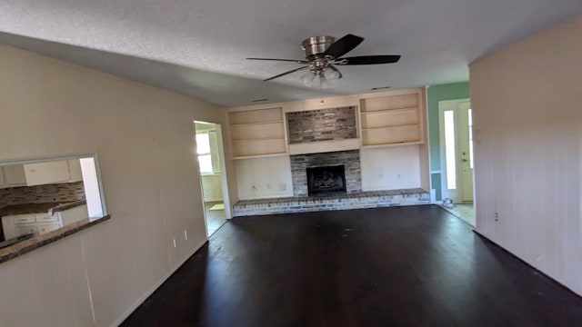 unfurnished living room with a fireplace with raised hearth, a ceiling fan, built in shelves, and dark wood-style flooring