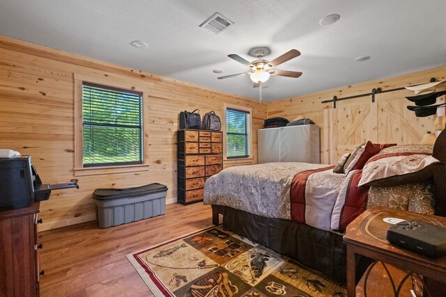 bedroom with a barn door, wooden walls, visible vents, light wood-type flooring, and ceiling fan