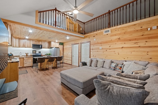 living area with a towering ceiling, wooden walls, visible vents, a ceiling fan, and light wood-style flooring