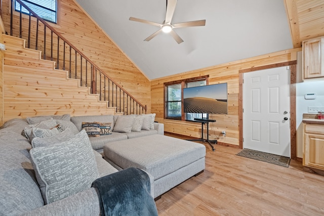 living room with light wood-type flooring, a ceiling fan, wood walls, stairway, and high vaulted ceiling