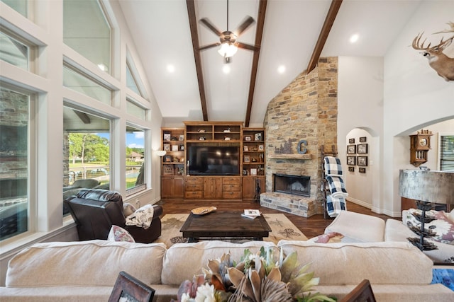 living area with beam ceiling, wood finished floors, ceiling fan, a stone fireplace, and high vaulted ceiling