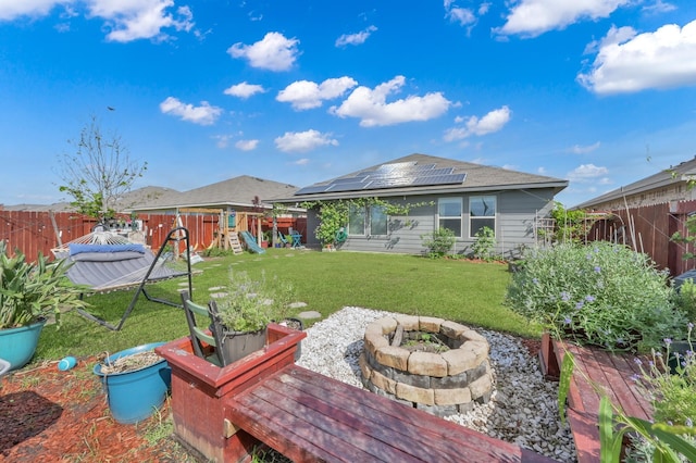 back of house with roof mounted solar panels, a playground, a fenced backyard, a lawn, and an outdoor fire pit