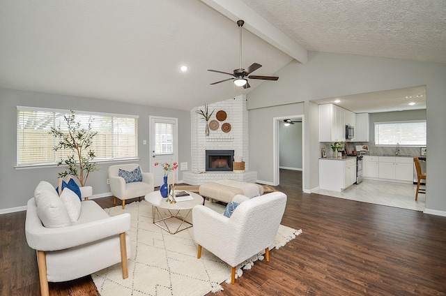 living area with a ceiling fan, vaulted ceiling with beams, light wood-style floors, baseboards, and a brick fireplace