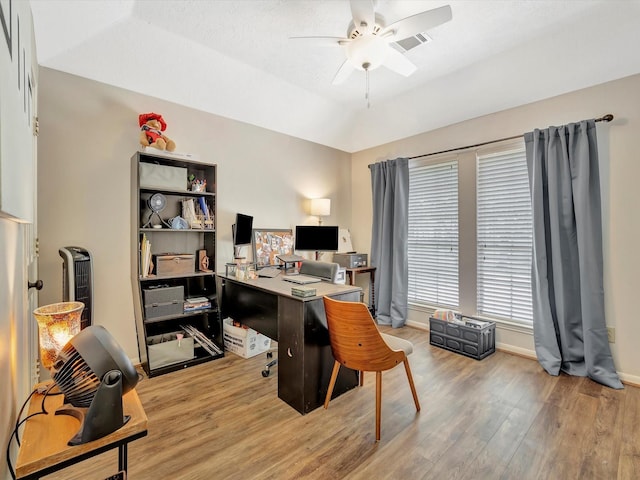 home office featuring light wood finished floors, vaulted ceiling, visible vents, baseboards, and ceiling fan