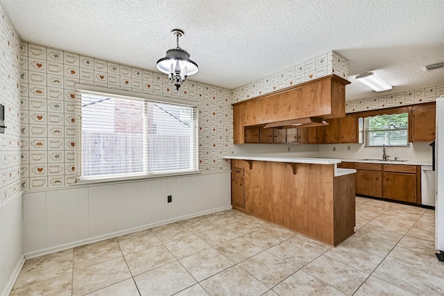 kitchen with brown cabinetry, a textured ceiling, a sink, light countertops, and wallpapered walls