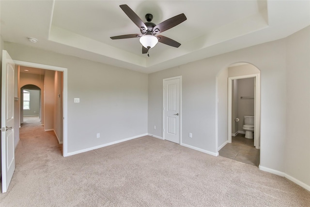 unfurnished bedroom featuring a raised ceiling, light carpet, connected bathroom, arched walkways, and baseboards