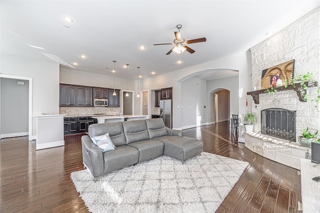 living area with ceiling fan, a stone fireplace, arched walkways, baseboards, and dark wood-style flooring