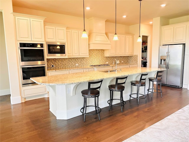 kitchen with stainless steel appliances, a sink, a center island with sink, decorative backsplash, and premium range hood