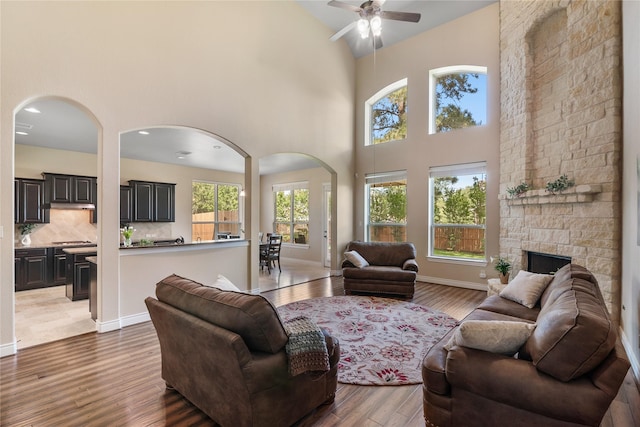 living room featuring a stone fireplace, wood finished floors, ceiling fan, arched walkways, and a high ceiling