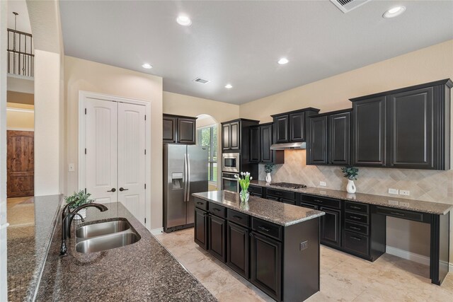 kitchen featuring arched walkways, a sink, dark cabinetry, a kitchen island, and appliances with stainless steel finishes