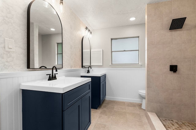 bathroom with wainscoting, a sink, a textured ceiling, two vanities, and tiled shower