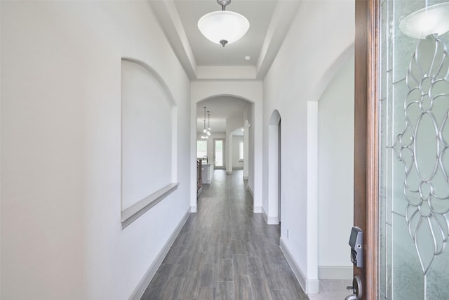 corridor with wood finished floors, baseboards, arched walkways, and a tray ceiling