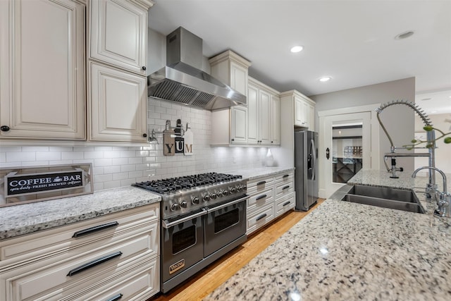 kitchen with light wood-style floors, wall chimney range hood, light stone counters, a sink, and appliances with stainless steel finishes