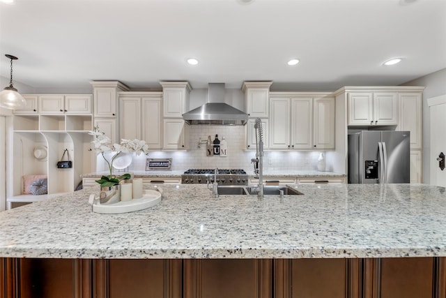 kitchen with a spacious island, wall chimney exhaust hood, stainless steel fridge with ice dispenser, light stone counters, and a sink