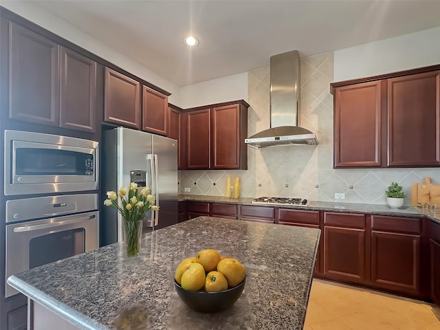 kitchen with wall chimney range hood, appliances with stainless steel finishes, dark stone counters, and reddish brown cabinets