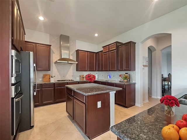 kitchen with wall chimney range hood, dark stone counters, arched walkways, backsplash, and stainless steel appliances