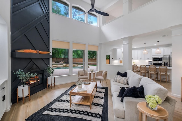 living area featuring ceiling fan, a glass covered fireplace, light wood-type flooring, a towering ceiling, and ornate columns